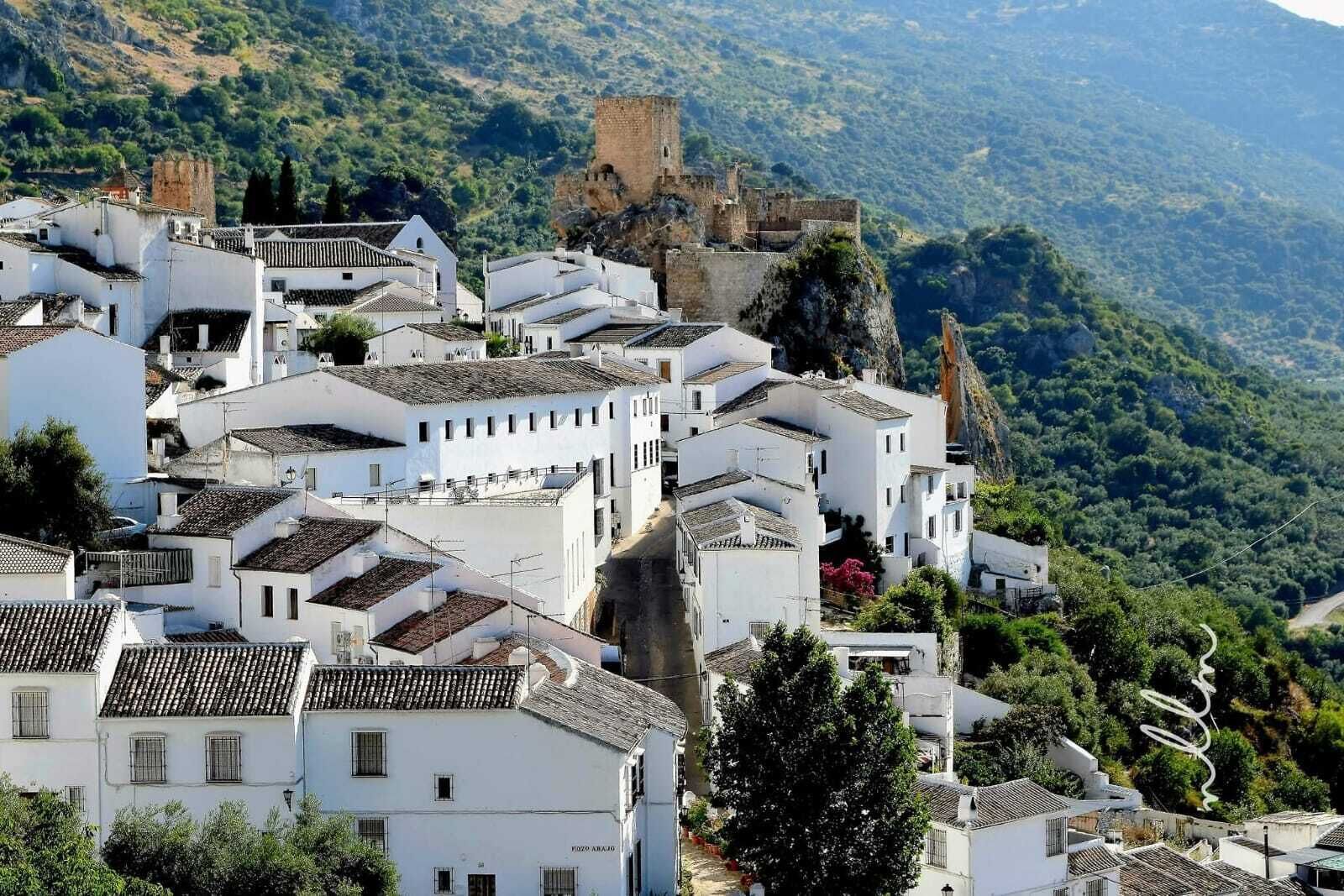 A white-walled village with terracotta roofs, a prominent castle on a hill, and a winding road.