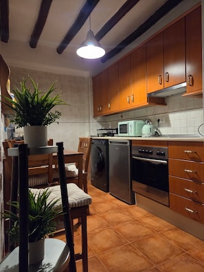 A kitchen with wooden cabinets, a washing machine, a microwave, and a dining area with chairs and a table.