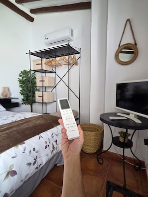 Een hand houdt een afstandsbediening vast, een bed met een bloemenprint, een televisie op een standaard, een plant en een metalen plank met dozen en kledinghangers.