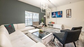 A modern living room with a white sofa, a glass coffee table, and a gray armchair. There are framed pictures on the wall and a plant on a side table. - Cozy Home in Amazing Resort for Perfect Vacations (Davenport)