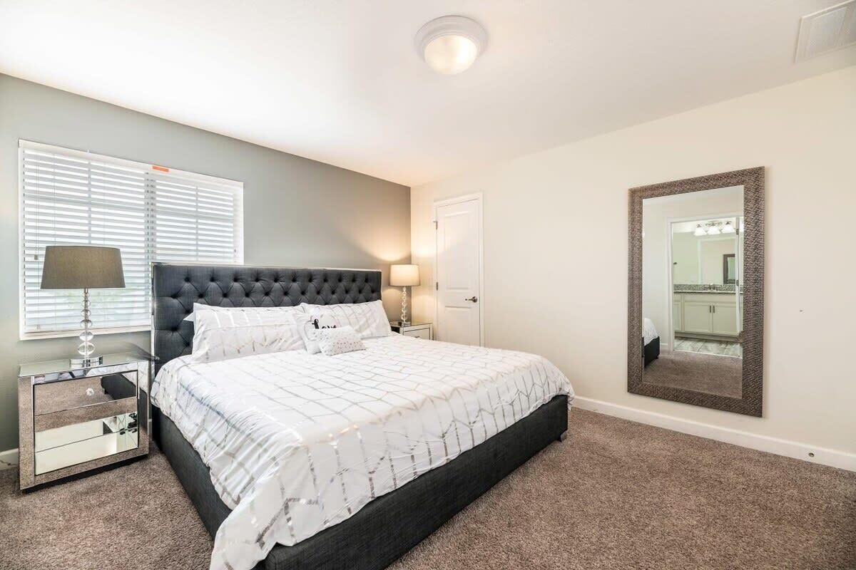 A bedroom with a large bed, a nightstand, a lamp, a mirror, and a window with blinds.
