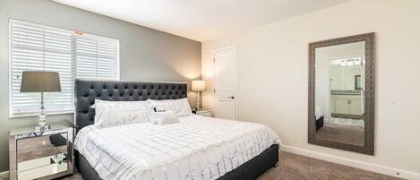 A bedroom with a large bed, a nightstand, a lamp, a mirror, and a window with blinds.