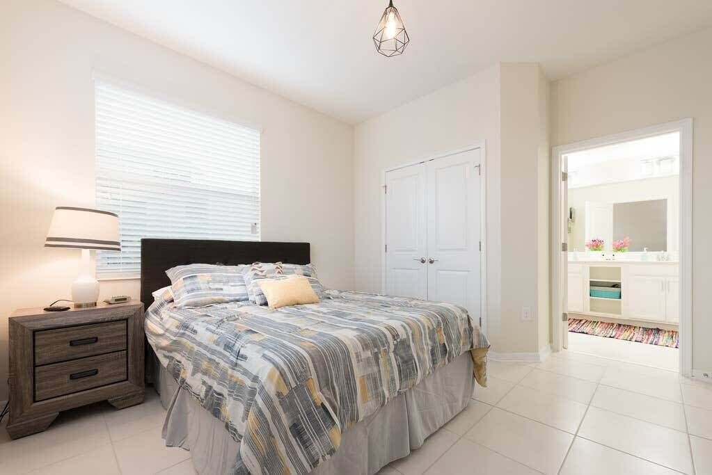 Un dormitorio con cama, mesita de noche, lámpara y una puerta que da a un baño.