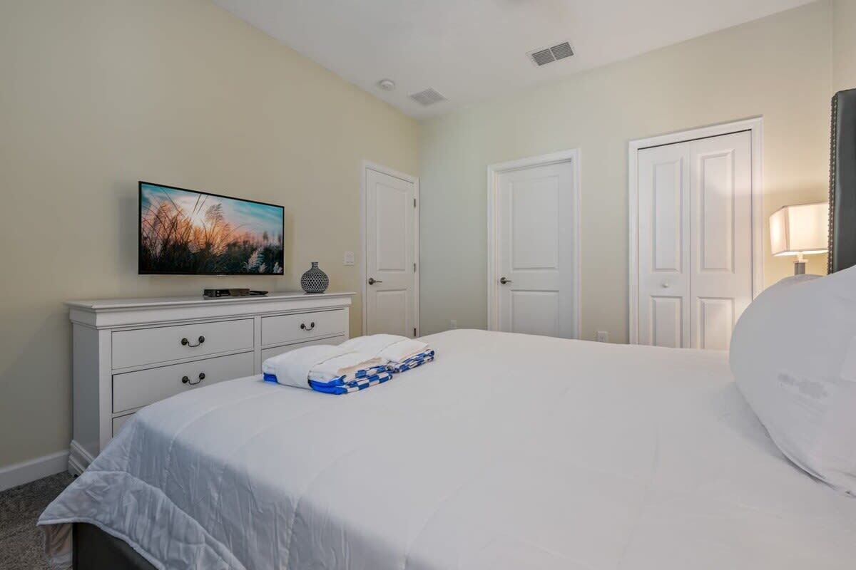 A neatly arranged bedroom with a bed, a dresser, a television, and two doors.
