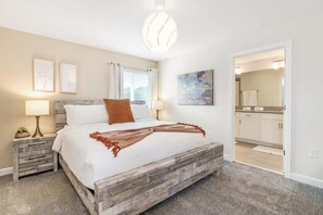 A bedroom with a wooden bed, two bedside tables, a nightstand, a modern light fixture, and a view into the bathroom. - Disney Dream Home, Prime Location, Private Pool (Kissimmee)