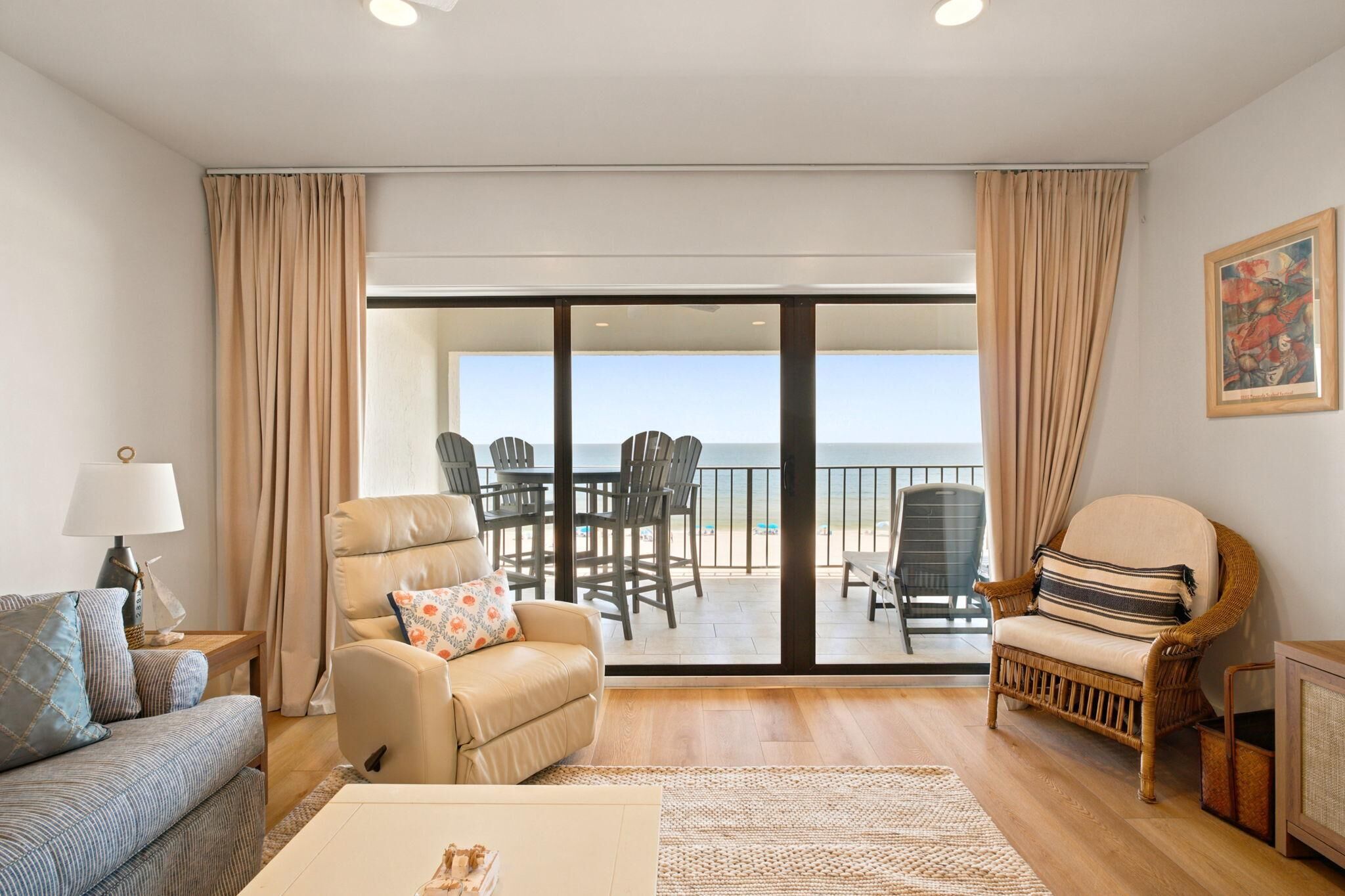 A living room with a sofa, armchair, and a dining table. There is a balcony with chairs and a view of the sea.