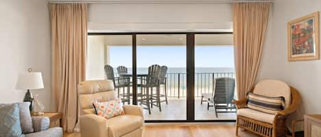Un salon comprenant un canapé, un fauteuil et une table à manger. Il y a un balcon avec des chaises et une vue sur la mer.