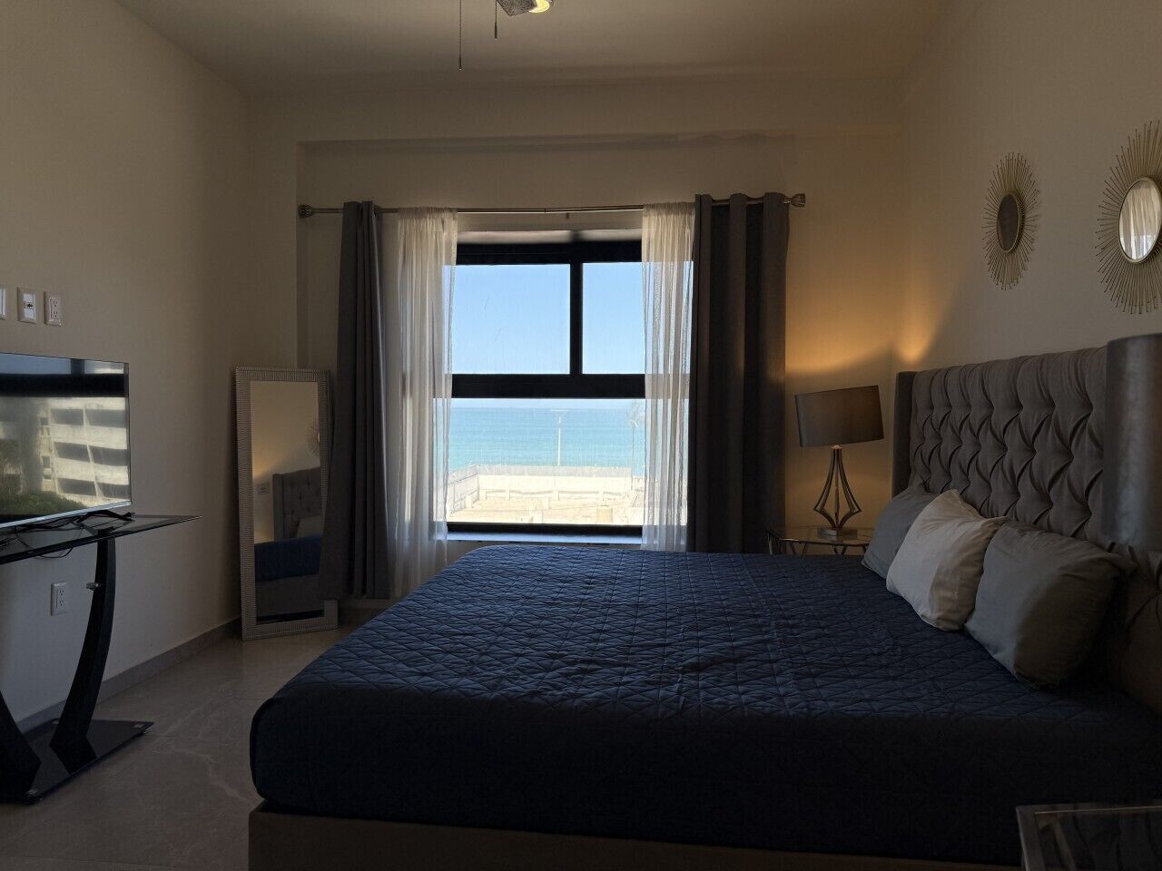 A hotel room with a bed, a desk, a chair, a lamp, and a view of the beach.