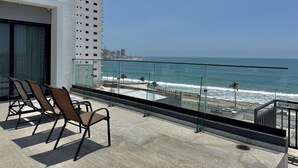 A balcony with chairs overlooking the ocean, a glass railing, and a view of the beach and cityscape. - Oceanview Condo in Palos Prietos | Pool + 1 Block to Beach (Mazatlán)