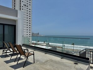 A balcony with chairs overlooking the ocean, a glass railing, and a view of the beach and cityscape. - Oceanview Condo in Palos Prietos | Pool + 1 Block to Beach (Mazatlán)