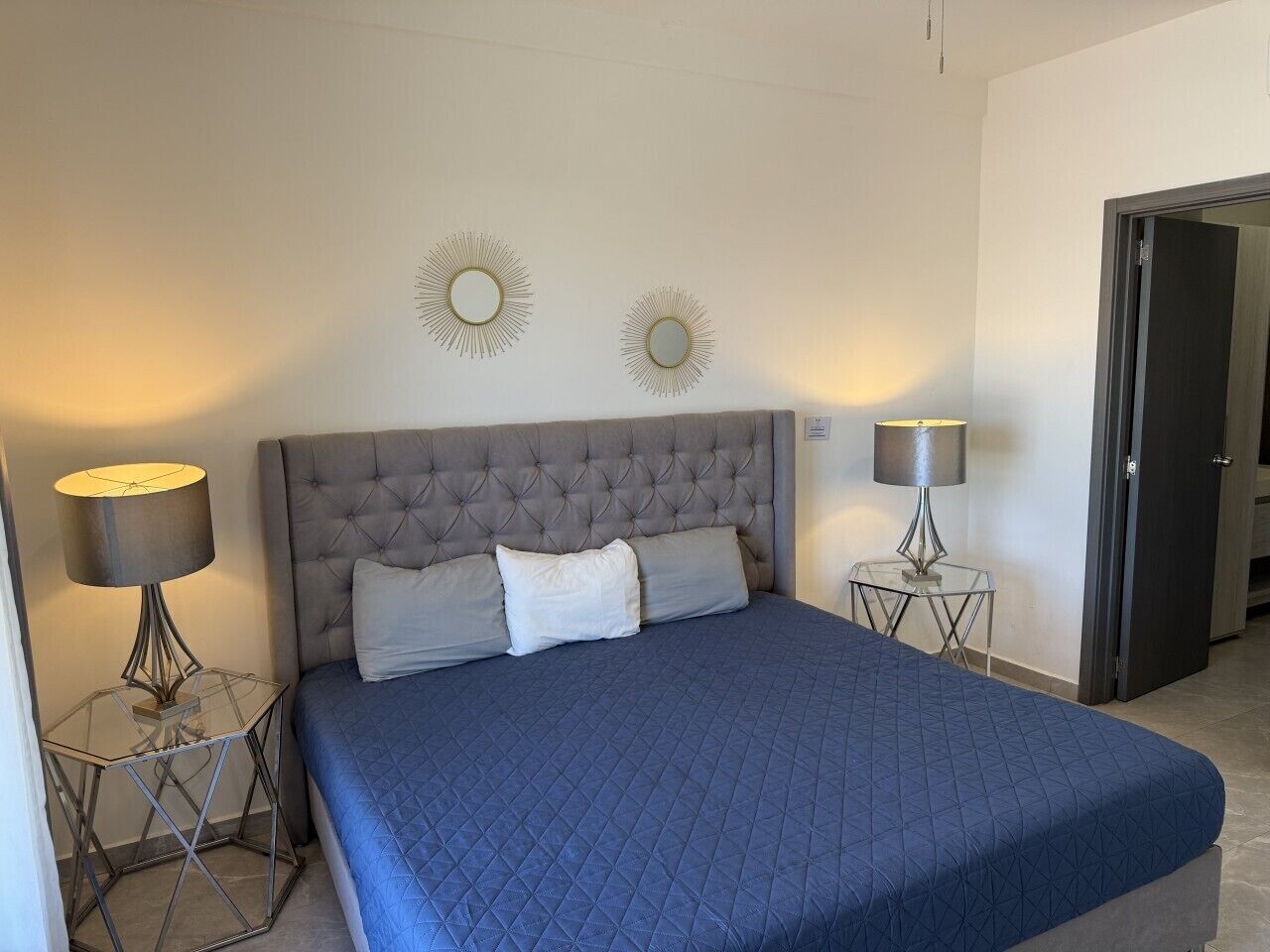 A bedroom with a tufted headboard, a large bed with a quilted blue bedspread, two bedside lamps, and two wall-mounted mirrors.
