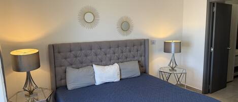 A bedroom with a tufted headboard, a large bed with a quilted blue bedspread, two bedside lamps, and two wall-mounted mirrors.