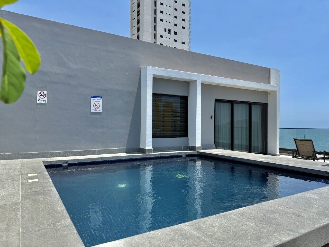 Une piscine, un bâtiment moderne, un panneau indiquant qu’il est interdit de fumer et une vue sur l’océan.