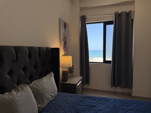 A bedroom with a bed, a nightstand, a lamp, a painting, and a window with curtains. - Oceanview Condo in Palos Prietos | Pool + 1 Block to Beach (Mazatlán)