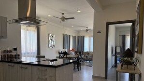A modern kitchen with a central island, dining area with chairs, and a living space with a sofa and curtains. - Oceanview Condo in Palos Prietos | Pool + 1 Block to Beach (Mazatlán)