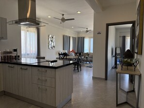 A modern kitchen with a central island, dining area with chairs, and a living space with a sofa and curtains. - Oceanview Condo in Palos Prietos | Pool + 1 Block to Beach (Mazatlán)