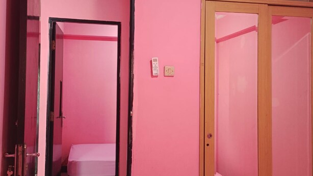 Una habitación con paredes rosas, un armario de madera, una cama y una puerta que da a otra estancia.