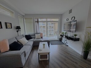 Ein modernes Wohnzimmer mit grauem Sofa, Couchtisch, Fernseher und einem Balkon mit Blick auf Gebäude.
