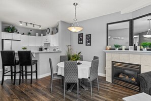 A modern kitchen with white cabinets, a dining area with a round table and grey chairs, and a fireplace with a mirror above it.