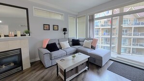 Un salon doté d’une cheminée, d’un canapé d’angle gris, d’une table basse et d’une grande fenêtre avec des stores.