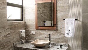 A modern bathroom with a granite countertop, a vessel sink, a wooden medicine cabinet, a towel rack, and a window. - Hotel Asís (San Juan de los Lagos)