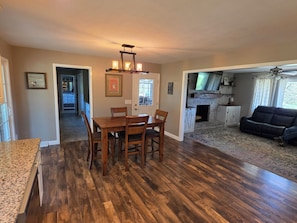 A dining area with a wooden table and chairs, a kitchen with a fireplace, and a living room with a sofa and a ceiling fan.