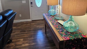 Una habitación con piso de madera, un sillón de cuero azul, una mesa con mantel colorido, dos lámparas y obras de arte en las paredes.