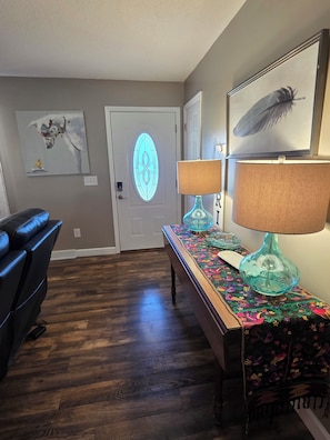 A room with a wooden floor, a blue leather chair, a table with a colorful tablecloth, two lamps, and artwork on the walls.