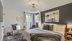 A bedroom with a large bed, a chandelier, a painting, and a view of the outdoors. - Inviting & Serene: A Book Lover’s Haven (Homewood)
