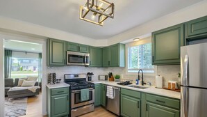 A modern kitchen with green cabinets, stainless steel appliances, and a dining area with a grey sofa. - Inviting & Serene: A Book Lover’s Haven (Homewood)