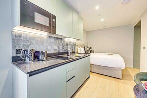 A modern kitchen with a built-in oven, a coffee maker, and a toaster. There is a bed with a white comforter and a bedside table with a lamp. - Superior studios at Farringdon by Dmontrio (London)