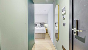 Un estrecho pasillo que conduce a un dormitorio con una cama, una mesita de noche, un espejo y una puerta con pomo.