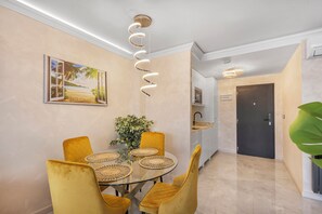 Un comedor con mesa de vidrio, sillas amarillas, un protector de mesa tejido, un cuadro en la pared y una lámpara con forma de espiral.