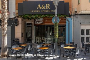 Un espacio al aire libre con mesas y sillas, un letrero que indica 'A&R Luxury Apartment' y un escaparate con la señalización 'CREPERIE PAISSERIE'.