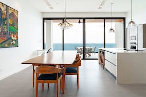 Eine moderne Küche mit einer großen Kochinsel, einem Essbereich mit Holztisch und Stühlen sowie Meerblick durch große Glasüren.