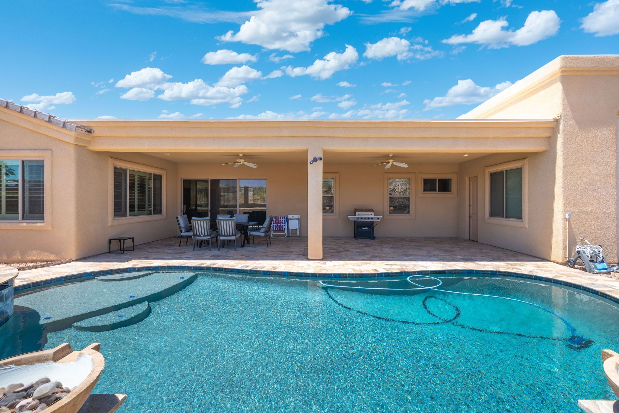 Una piscina con zona de patio, que incluye sillas y una mesa, y una sección cubierta con ventilador de techo.