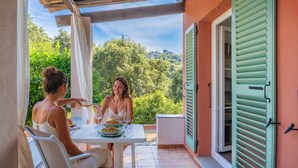 An outdoor dining area with a table set for two, a view of greenery and buildings in the background, and a building entrance with green shutters. - La Ty Kaz – Cocon entre mer, maquis et piscine en Costa Verde (Valle-di-Campoloro)