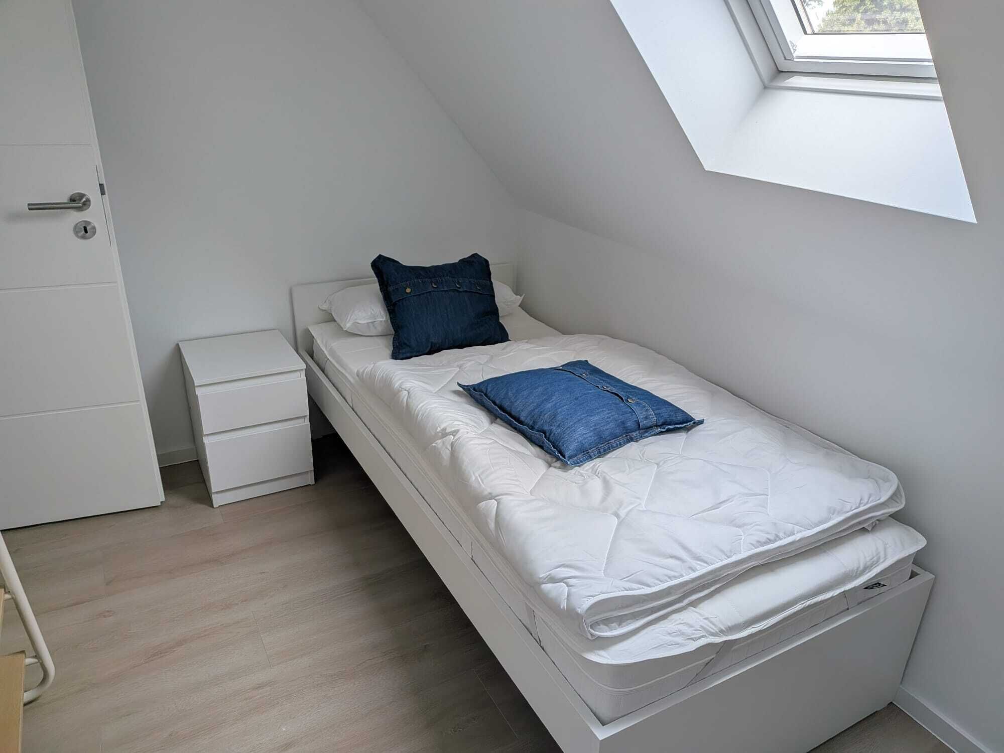 Ein Einzelbett mit weißer Bettwäsche und zwei blauen Kissen, ein kleiner Nachttisch und ein Fenster mit Blick auf Bäume.