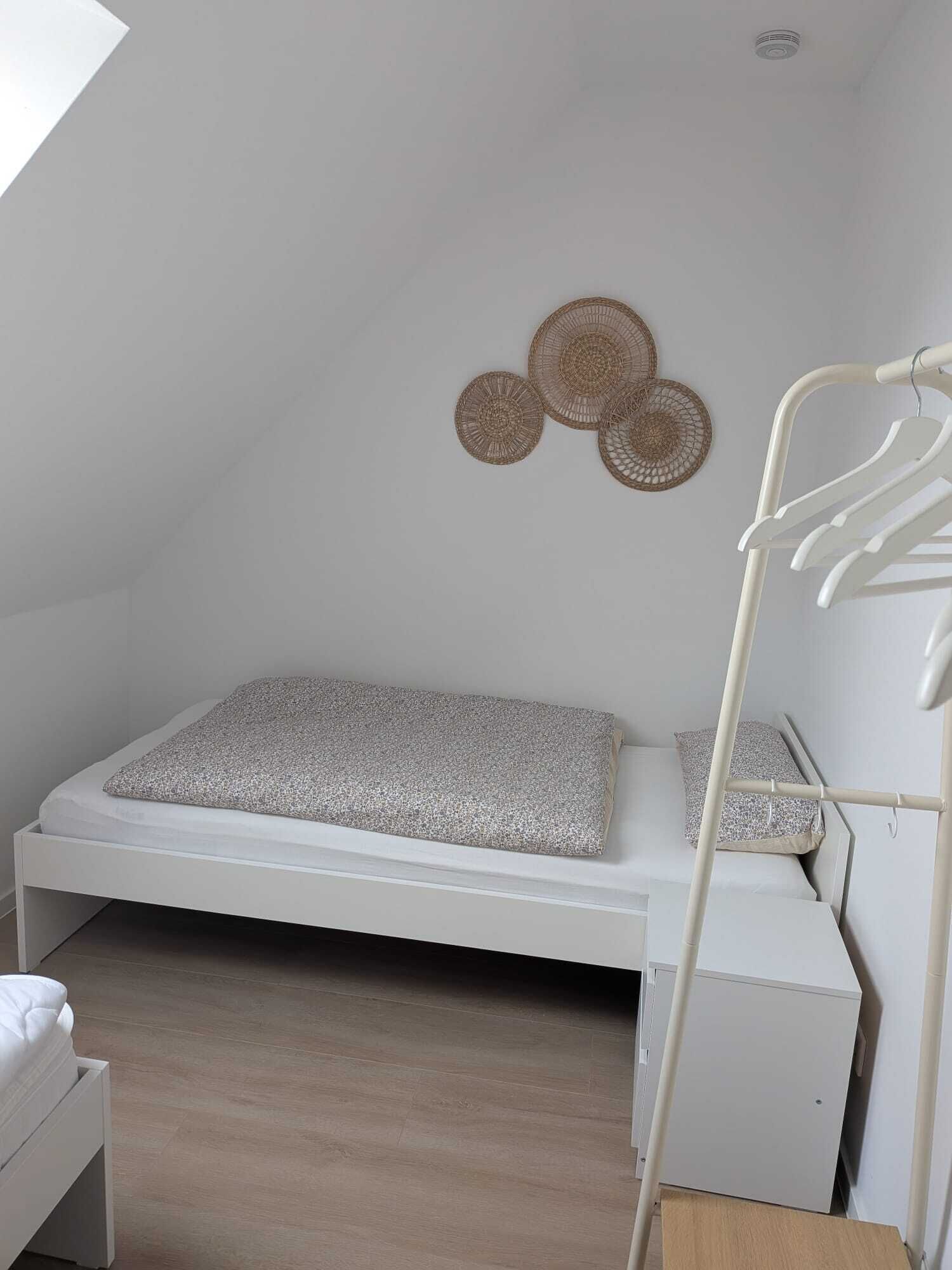 A small, white, single-bed room with a sloped ceiling, a wall-mounted drying rack, and a woven wall decoration.