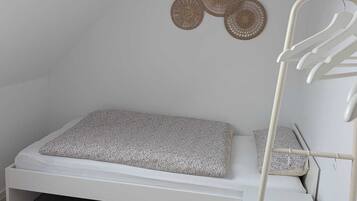 A small, white, single-bed room with a sloped ceiling, a wall-mounted drying rack, and a woven wall decoration.