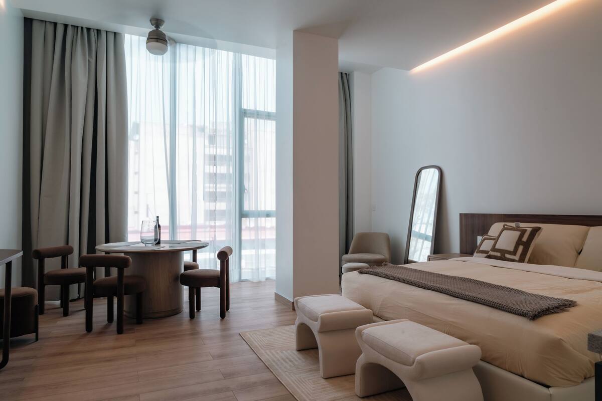A modern hotel room with a round dining table, four chairs, a large bed with a headboard, a bedside table, and a mirror.