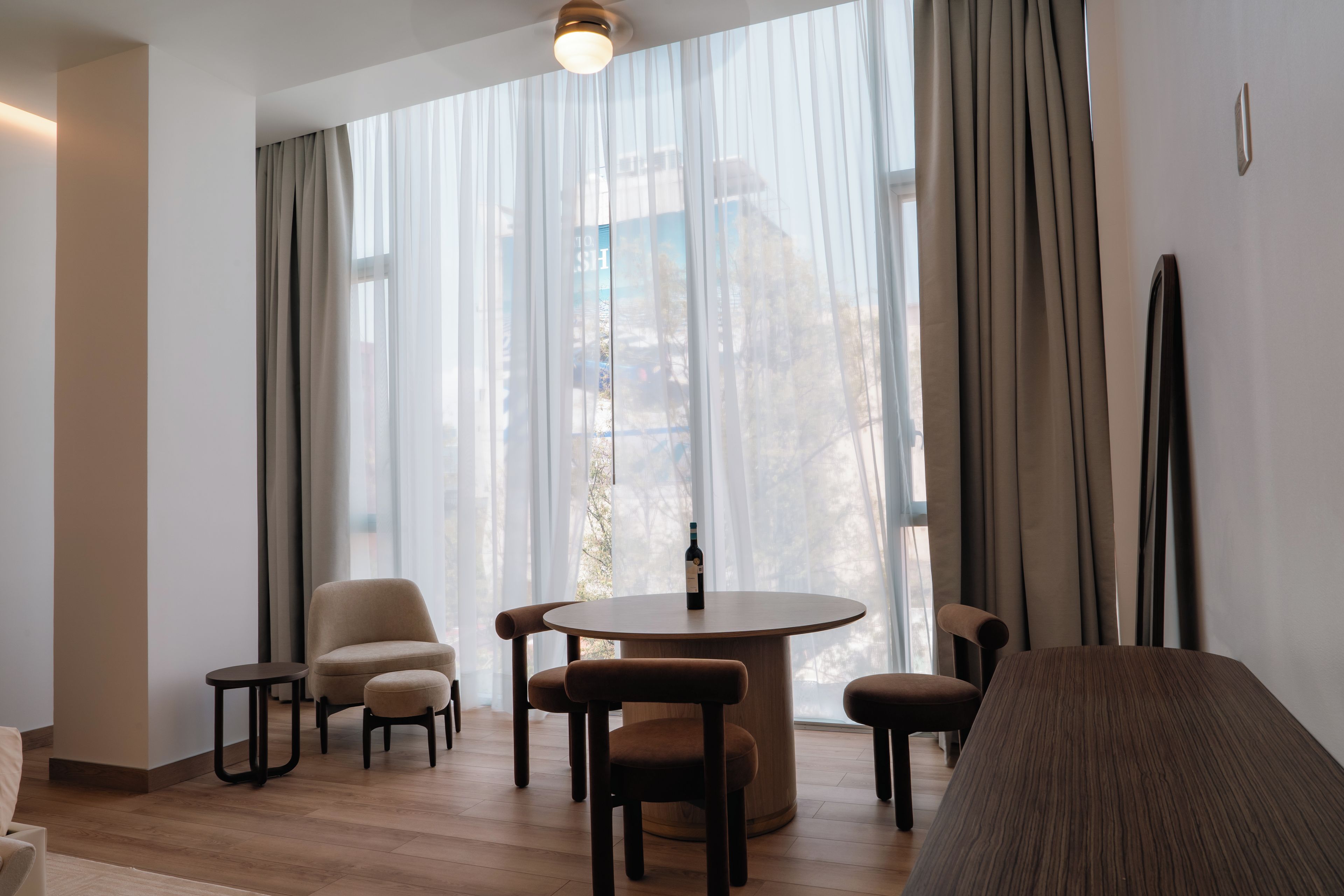 Quarto moderno com mesa redonda, quatro cadeiras, um sofá e uma janela grande com cortinas leves.