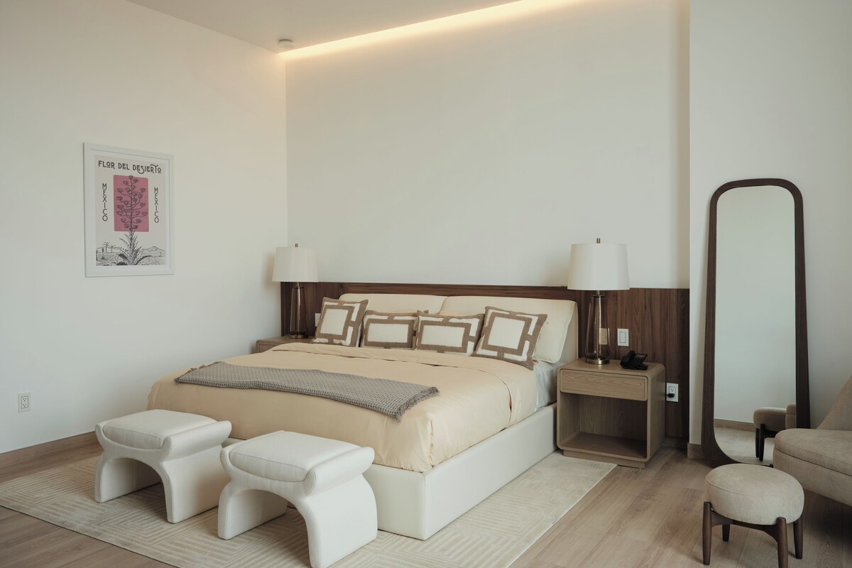 Ein modernes Schlafzimmer mit einem großen Bett, Nachttischlampen, einem Beistelltisch, einem Spiegel und einem kleinen Hocker.