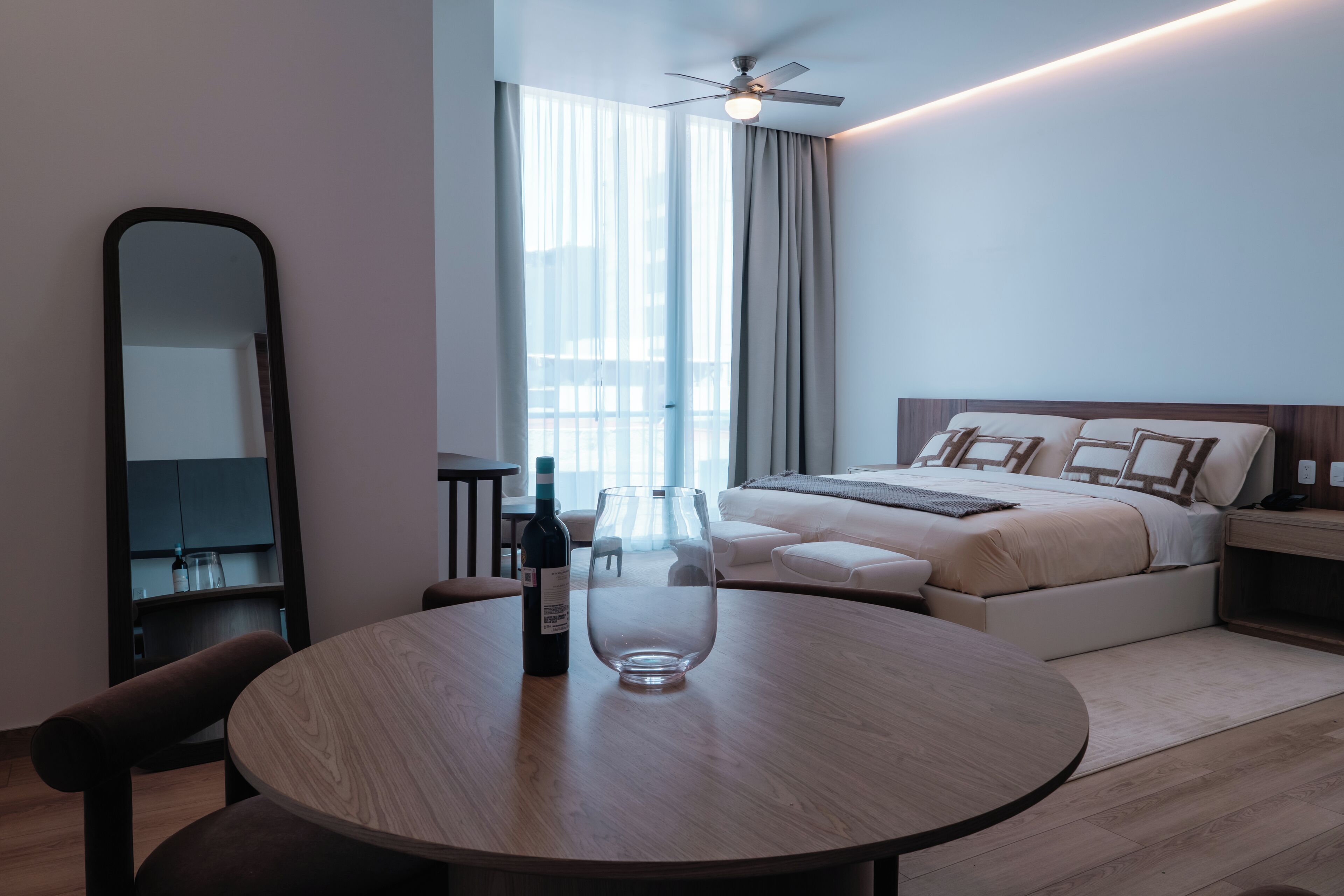 A modern hotel room with a round dining table, a minibar, a bed with a headboard, a ceiling fan, and a large window with curtains.