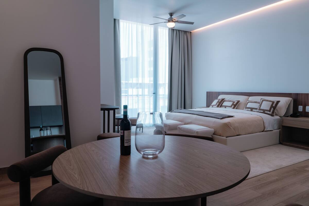 A modern hotel room with a round dining table, a minibar, a bed with a headboard, a ceiling fan, and a large window with curtains.
