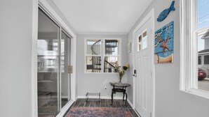 Un pasillo con una puerta de cristal que da a una sala de estar, una mesita con un florero y una obra de arte con motivos marinos en la pared.