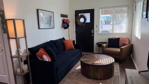 A living room with a dark blue sofa, a round coffee table, a small side table, a door with a wreath, and a window with blinds. - Charming 2 BR Little Oasis, with  pool, near Colorado River, SNOWBIRD FRIENLDY (Bullhead City)