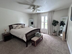A bedroom with a bed, a ceiling fan, a window with curtains, a mirror, and a potted plant.