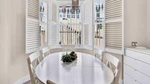 Een eetruimte met een ronde tafel, witte stoelen, een plantenarrangement in het midden en uitzicht door open witte luiken.