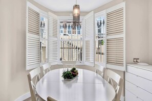 Ein Essbereich mit einem runden Tisch, weißen Stühlen, einer zentralen Pflanzenkomposition und Blick durch offene weiße Fensterläden.